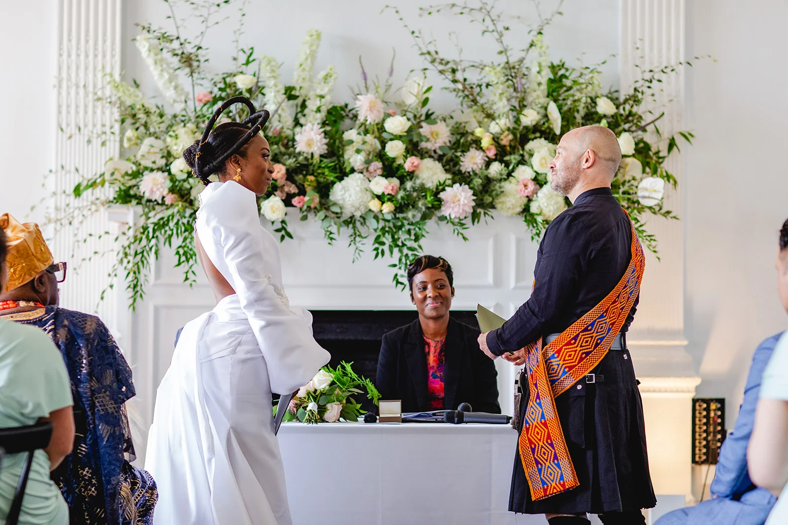 A bride in a modern white dress with a striking sculptural hairstyle stands facing the groom in a traditional kilt during their wedding ceremony.