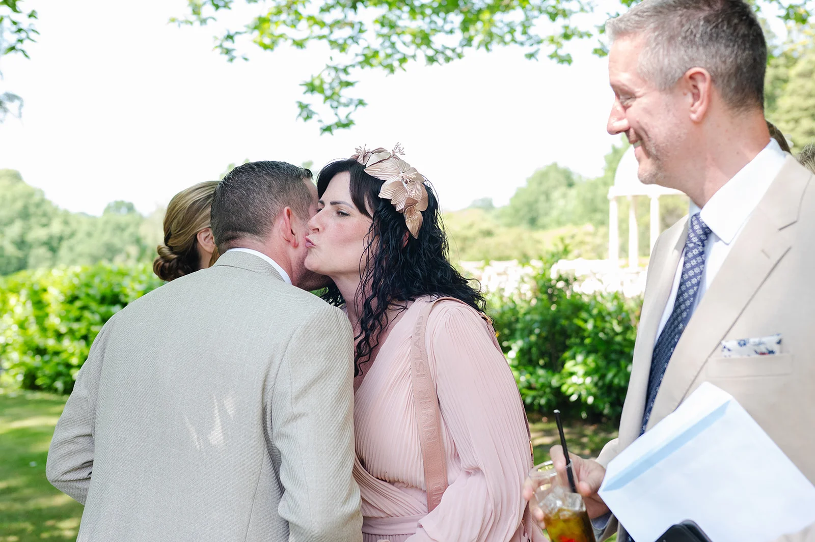 A guest wearing a pink dress and a floral headpiece greets a man with a kiss on the cheek during an outdoor wedding drinks reception.