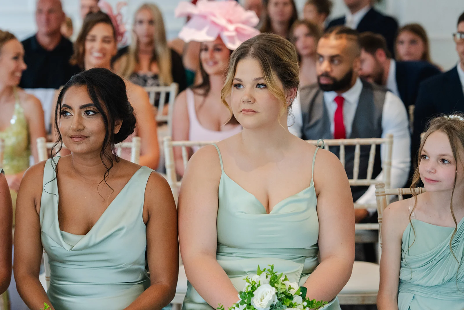 Three bridesmaids wearing sage green satin dresses with cowl necklines sit attentively together holding small white rose bouquets during a wedding ceremony.
