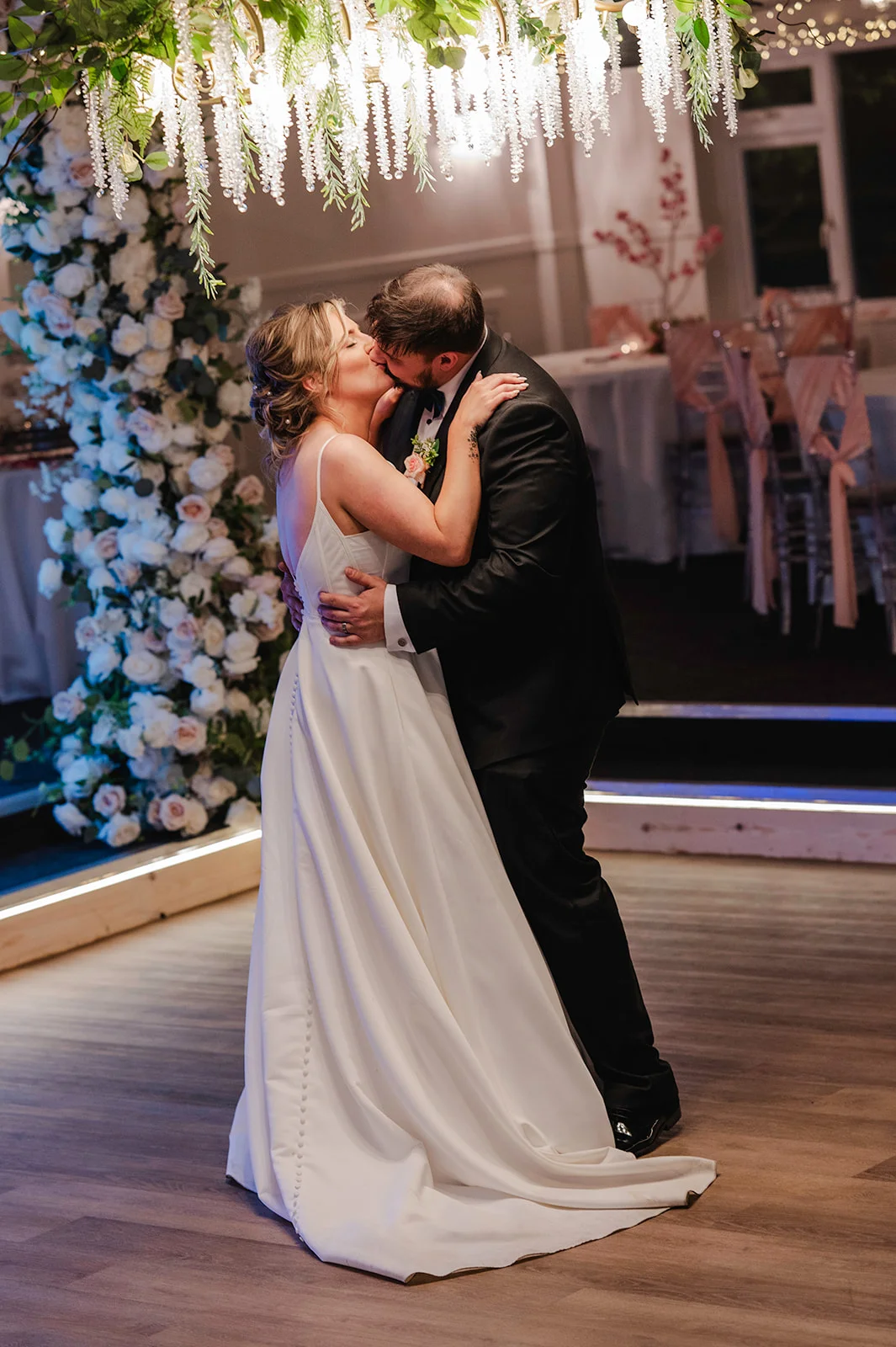 A bride in a sleek satin a-line gown and groom in a classic black tuxedo share a tender kiss beneath an illuminated crystal chandelier and a lush white floral arch.