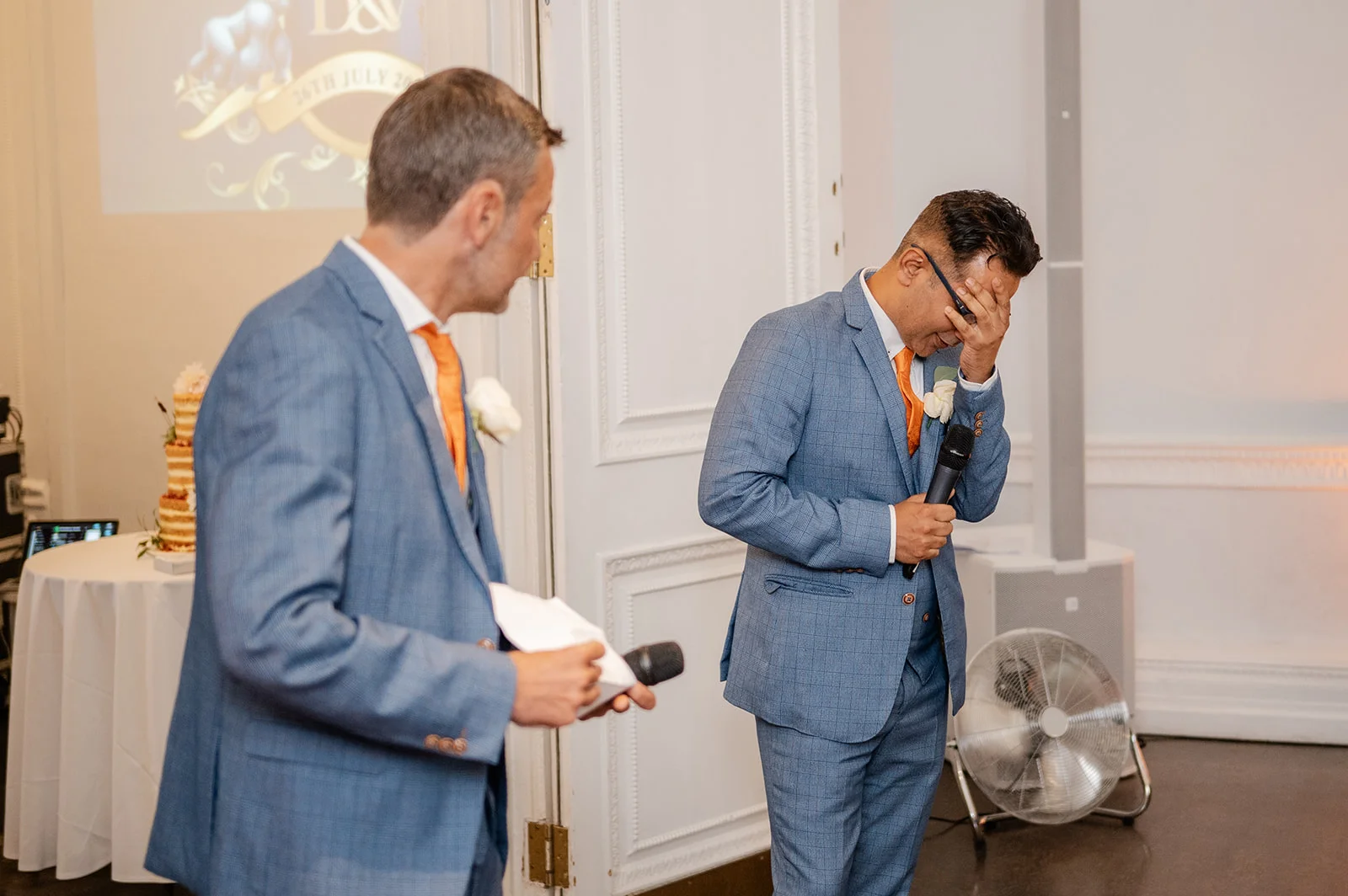 A groom covering his face in laughter or emotion while holding a microphone during the wedding speeches, standing next to a best man in a matching blue checked suit.