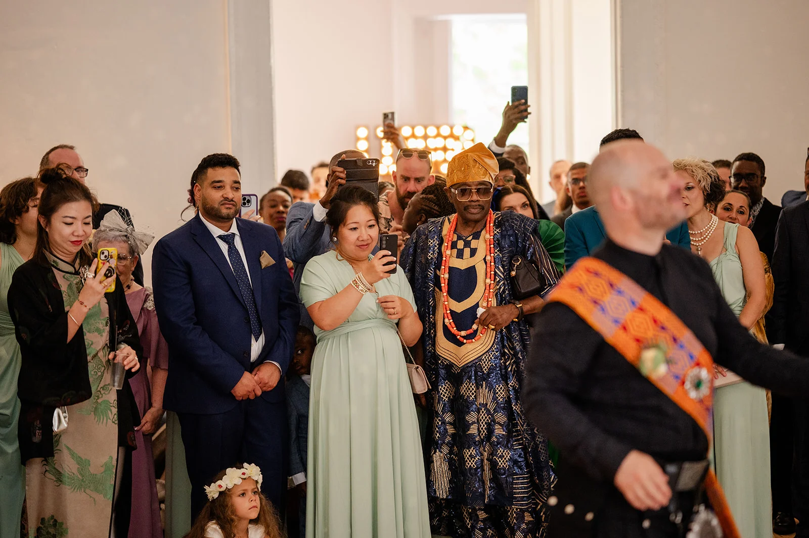 A candid group of wedding guests capture the ceremony on their phones while dressed in elegant formal wear and traditional attire.