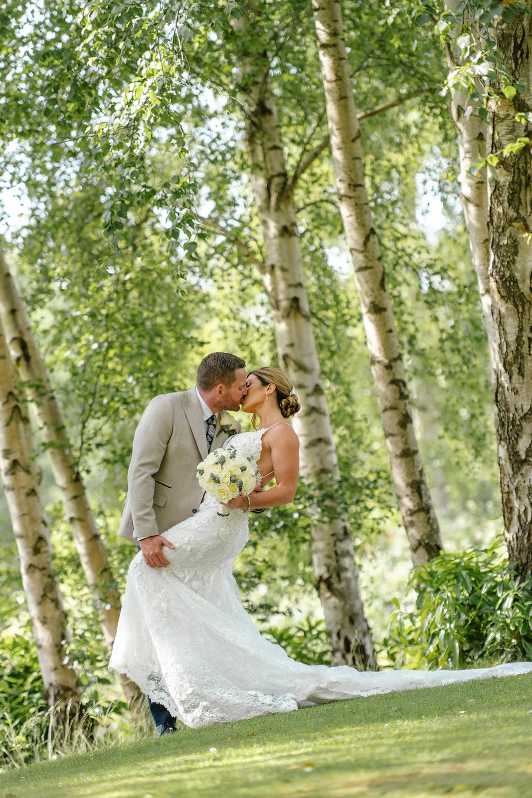 A newlywed couple shares a romantic dip kiss surrounded by tall birch trees in a sunlit woodland setting.