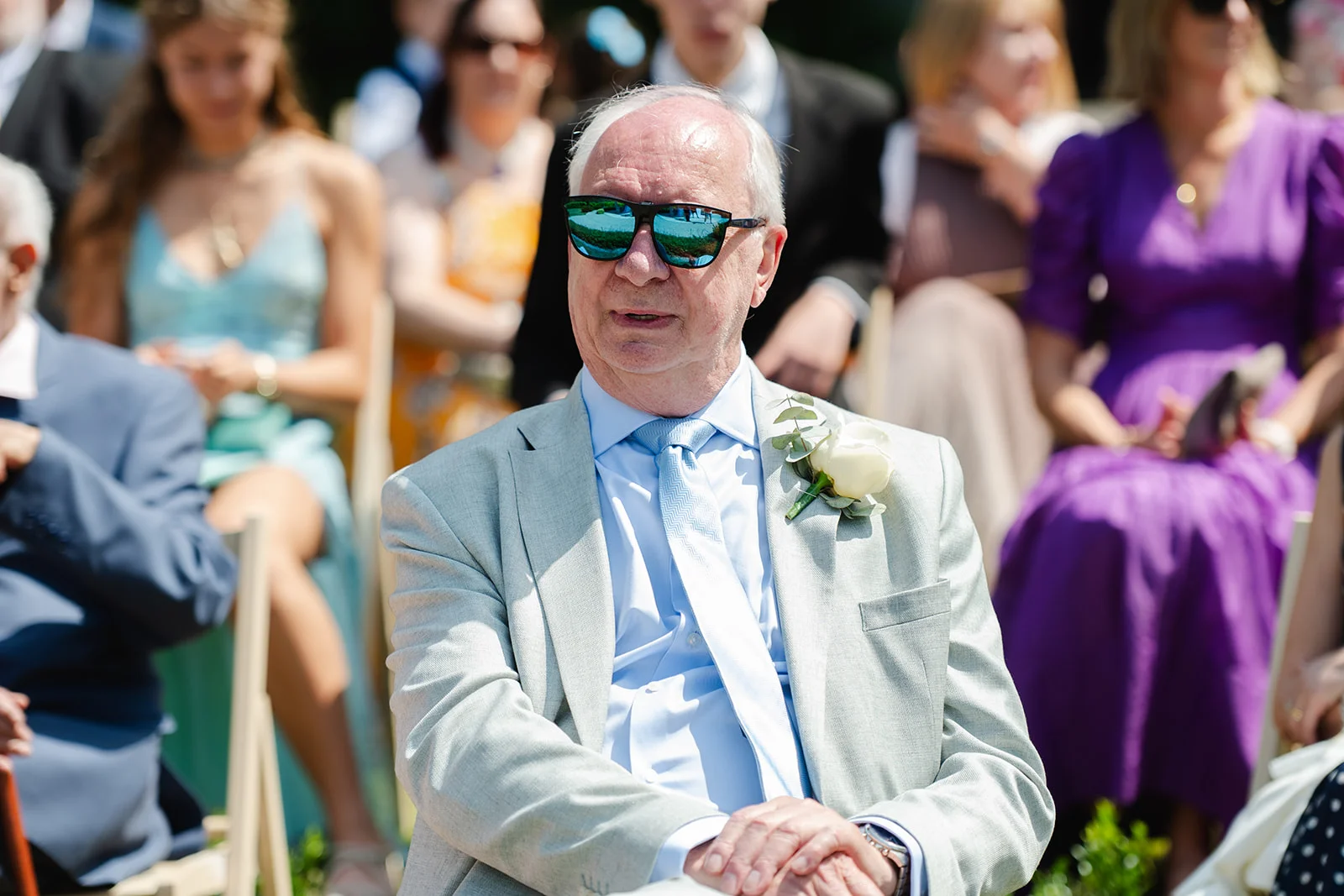 A distinguished male wedding guest wearing a light grey suit, blue shirt, matching tie, and reflective sunglasses sits among seated guests during an outdoor ceremony.