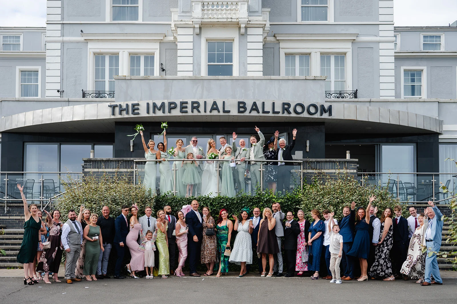 A large wedding party and guests wave and celebrate outside the grand entrance of The Imperial Ballroom hotel.