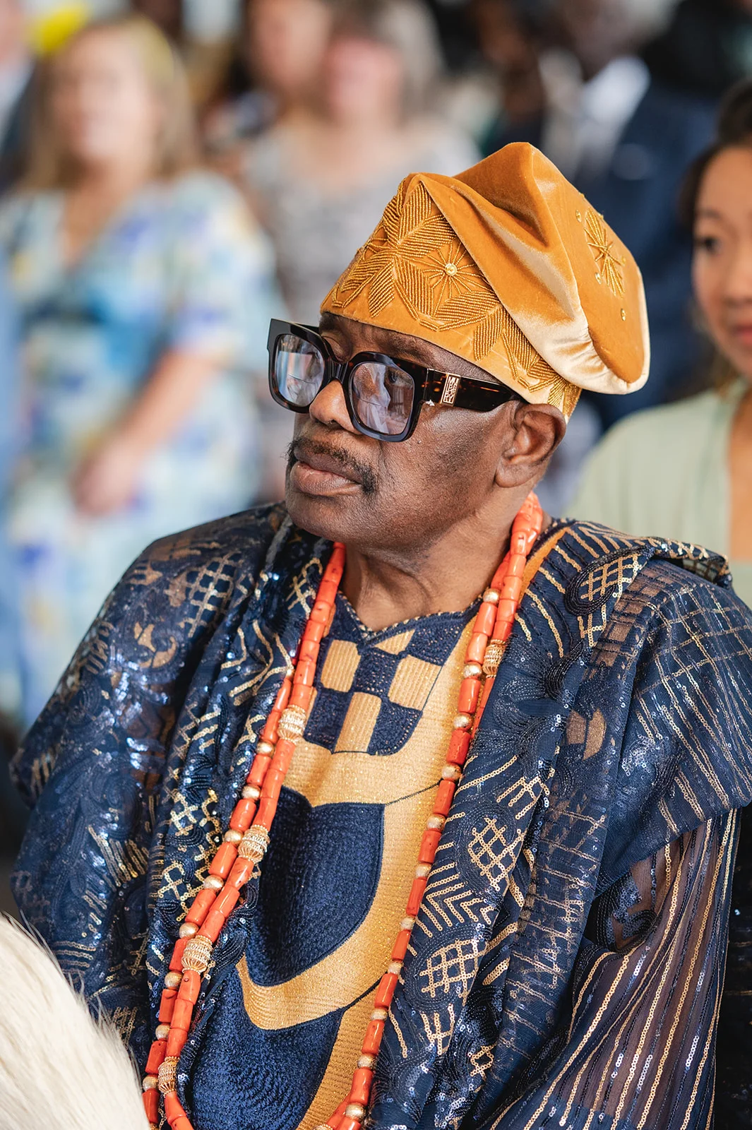 A guest at a wedding wearing traditional Nigerian attire with elaborate blue and gold patterned garments and bead necklaces.