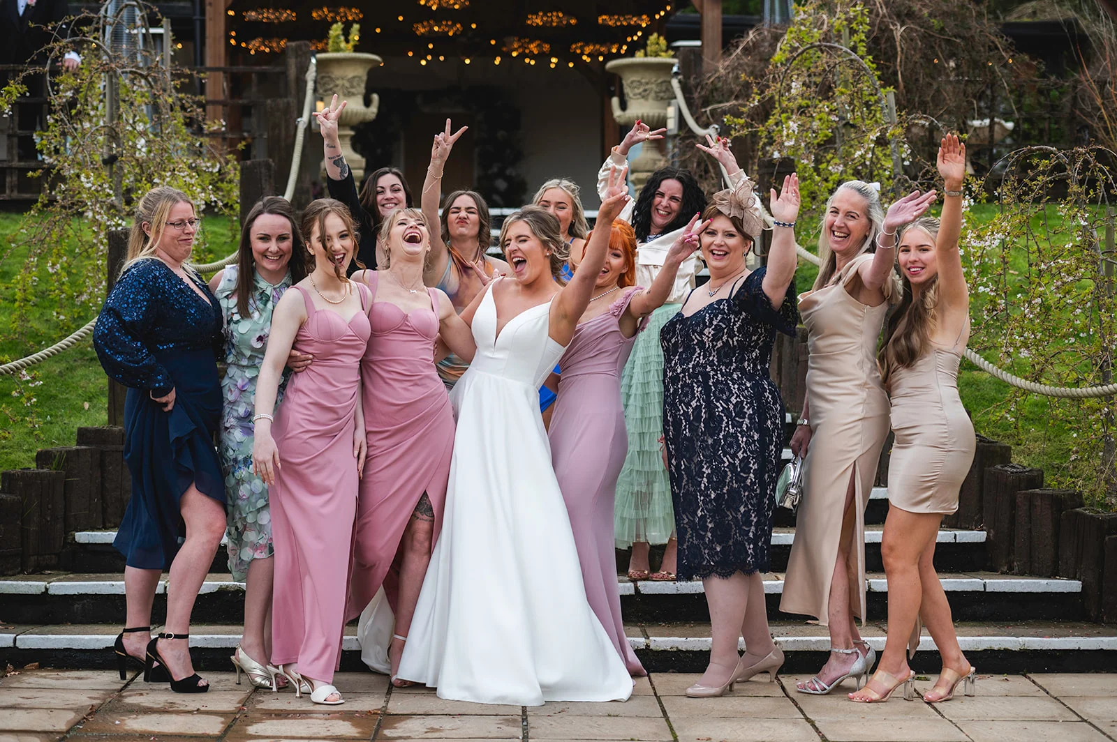 A joyful bride in a white a-line gown laughs excitedly while posing outside on steps with her diverse group of female friends and bridesmaids in mismatched dresses.