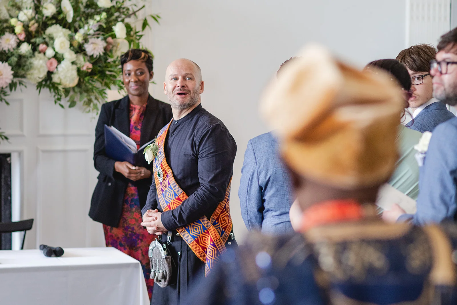 A smiling groom wearing a kilt and a colourful sash stands at the altar during a joyful humanist wedding ceremony.