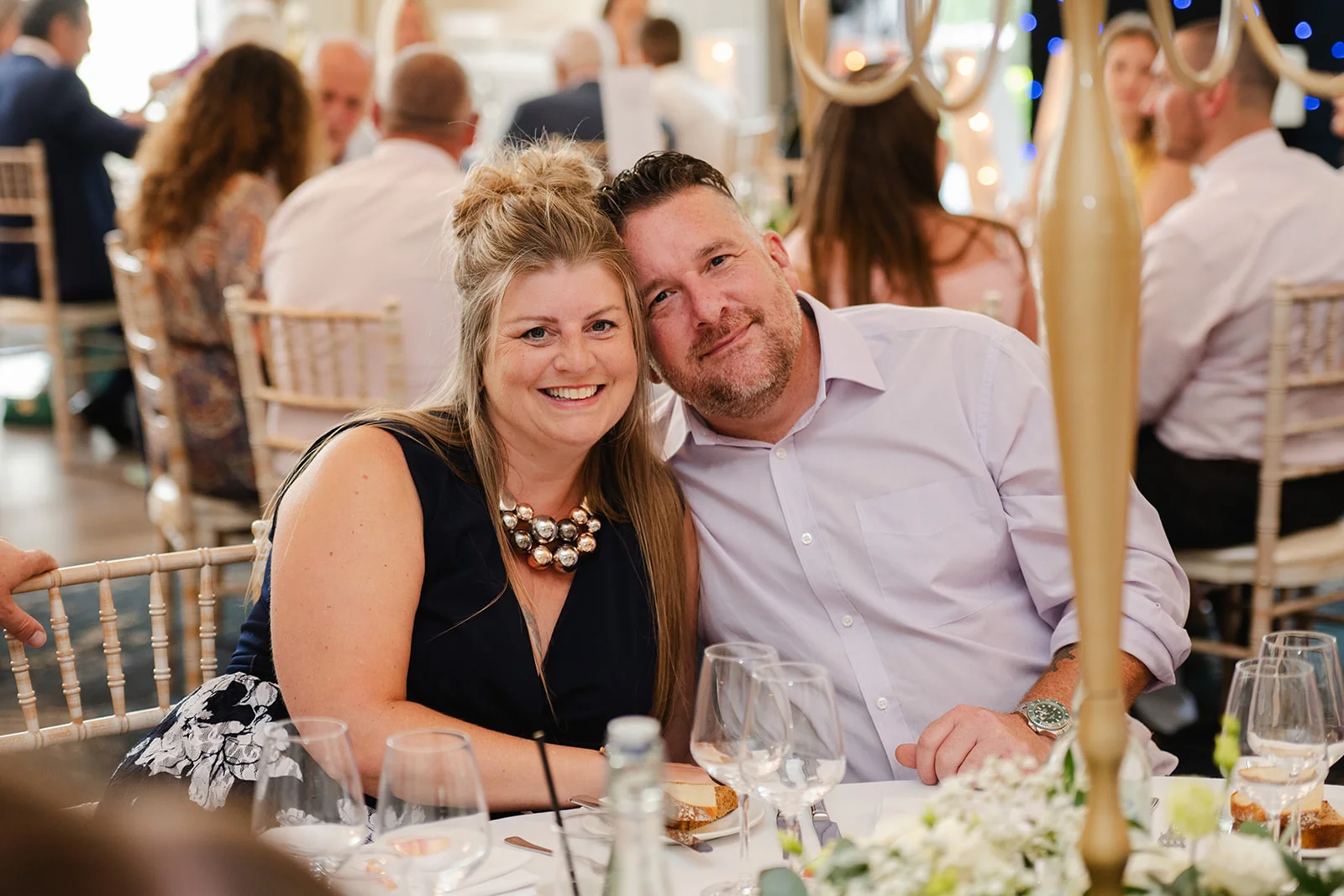 A smiling couple poses for a candid photograph seated at a round reception table during a wedding celebration.