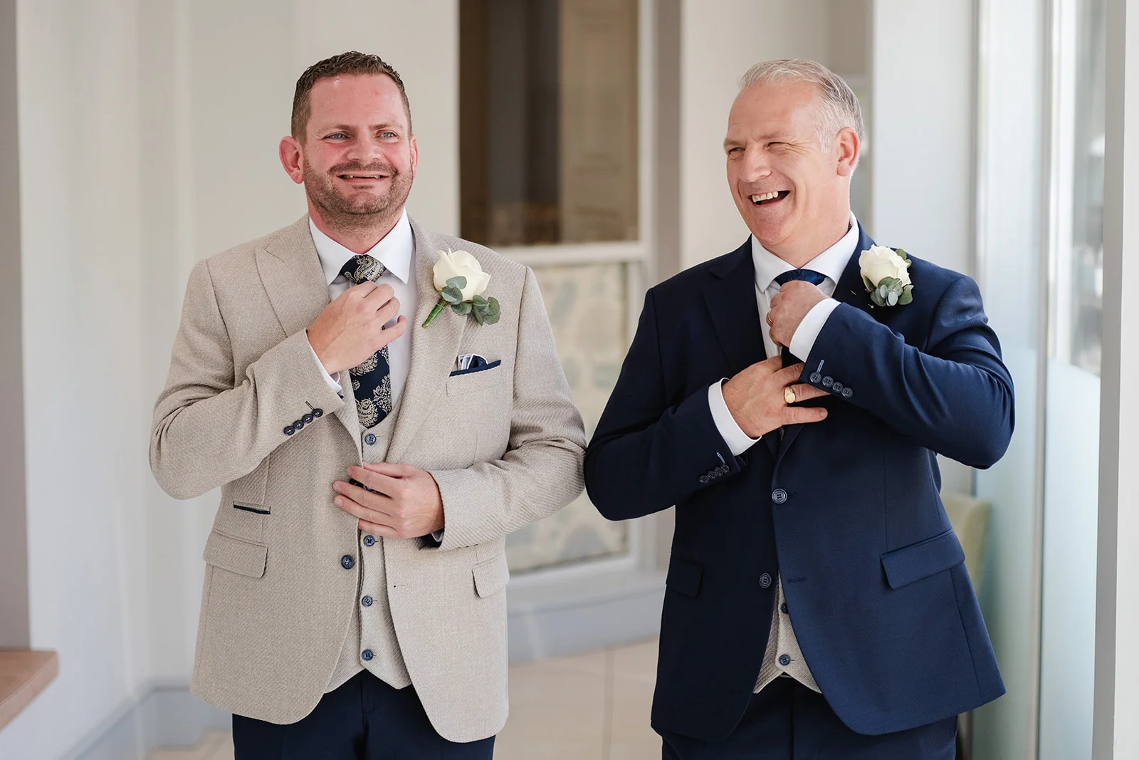 Two grooms laughing joyfully as they adjust their ties and jackets while getting ready on their wedding day.