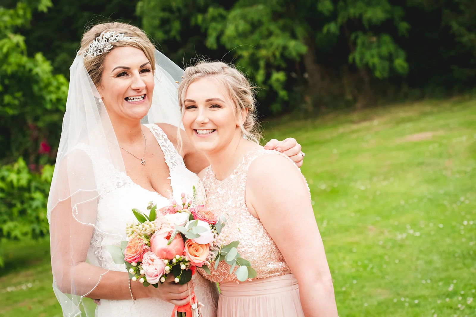 A joyful bride wearing a sparkling tiara and veil laughs heartily while holding a colourful pastel bouquet next to her bridesmaid in a shimmering pink dress outdoors in a lush green garden.