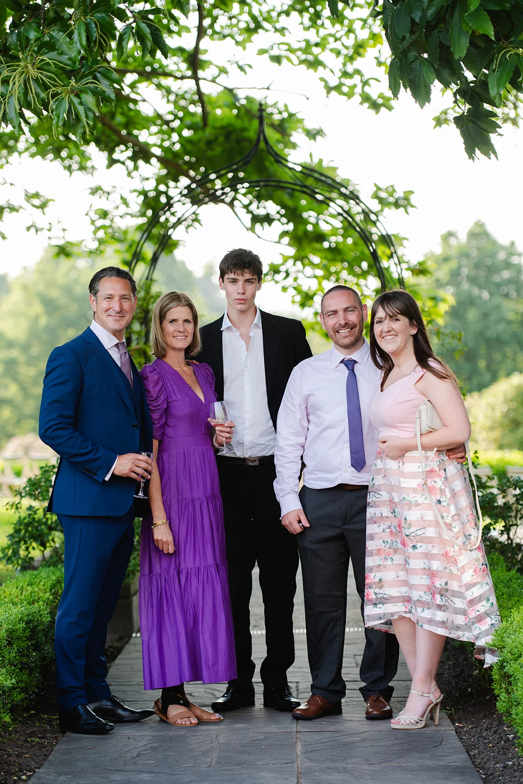 A group of five wedding guests pose together outdoors on a stone path beneath a metal archway in a manicured garden setting.