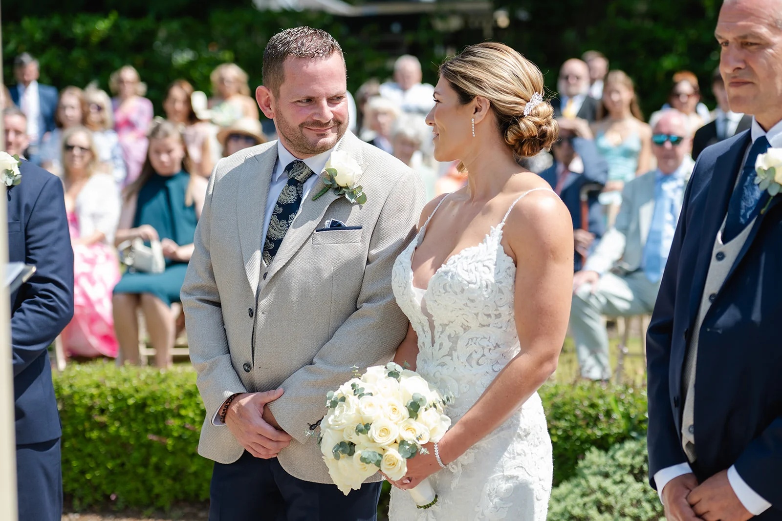 The bride and groom exchange a tender glance during their sunlit outdoor wedding ceremony, surrounded by seated guests.