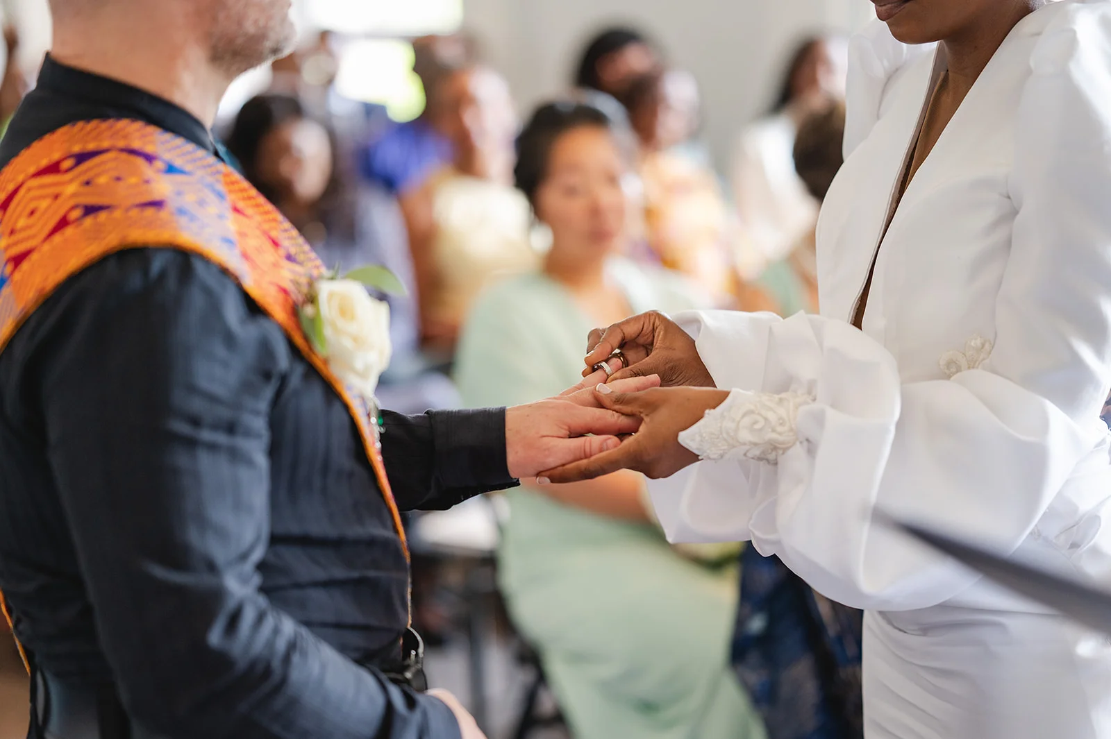 A close-up shot captures a bride placing a wedding ring onto the groom's finger during a heartfelt ceremony.