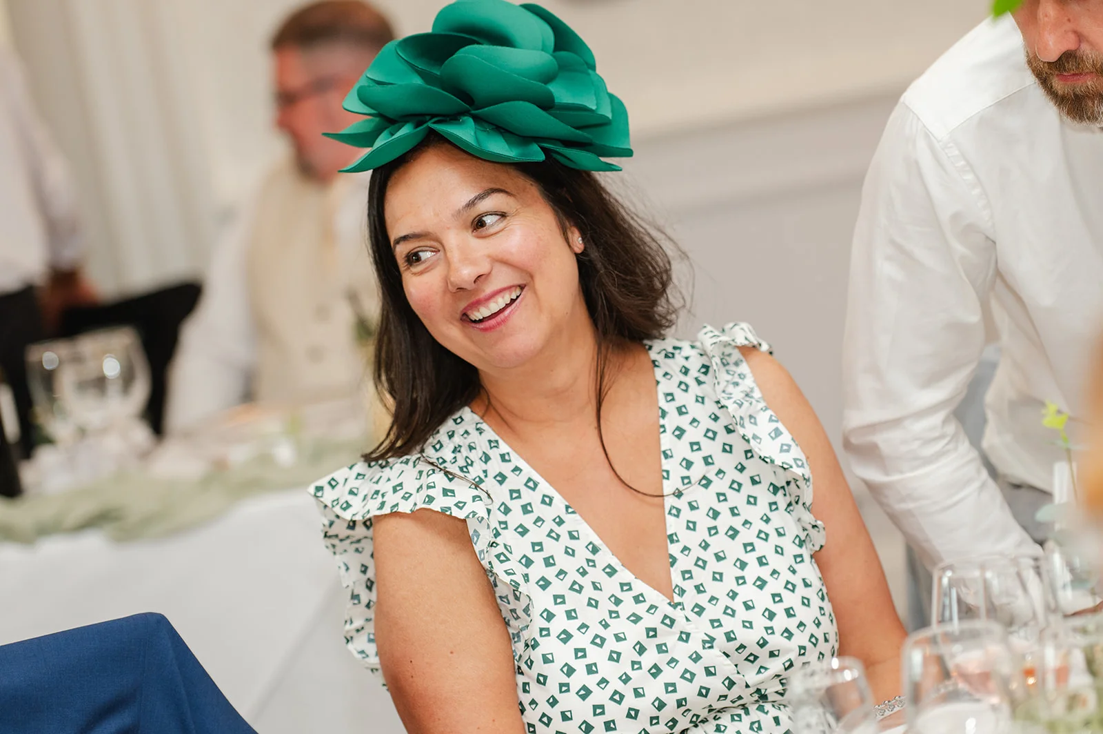 A smiling wedding guest wearing a vibrant green sculptural fascinator and a patterned green and white dress sits at a reception table.