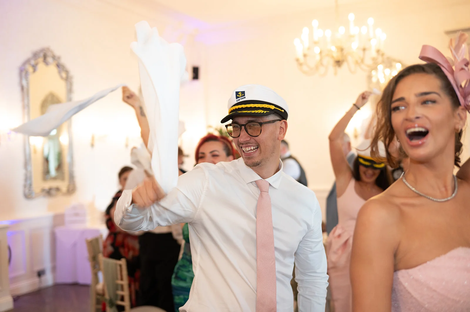 A joyful wedding guest wearing a captain's hat enthusiastically waves a white napkin in the air while celebrating at a lively reception.