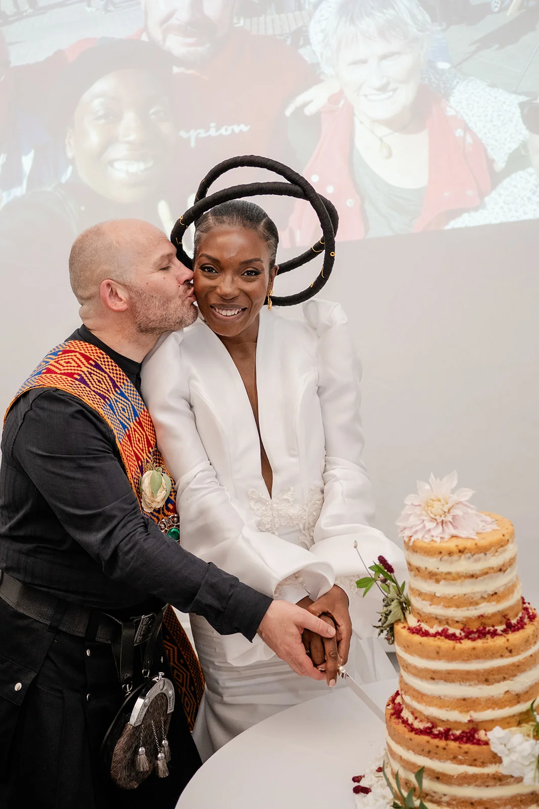 An interracial couple smiling while cutting a tiered semi-naked wedding cake, as the groom kisses the bride on the cheek.