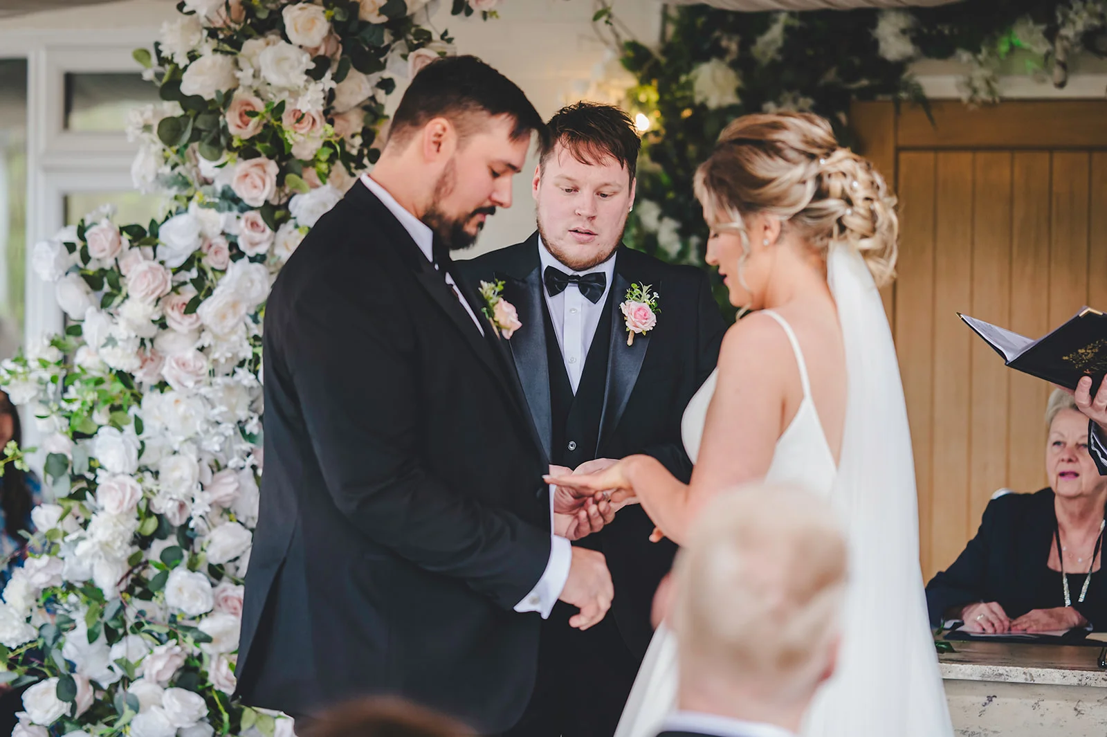 The groom places a wedding ring on the bride's finger during a heartfelt outdoor ceremony framed by a lush floral arch.