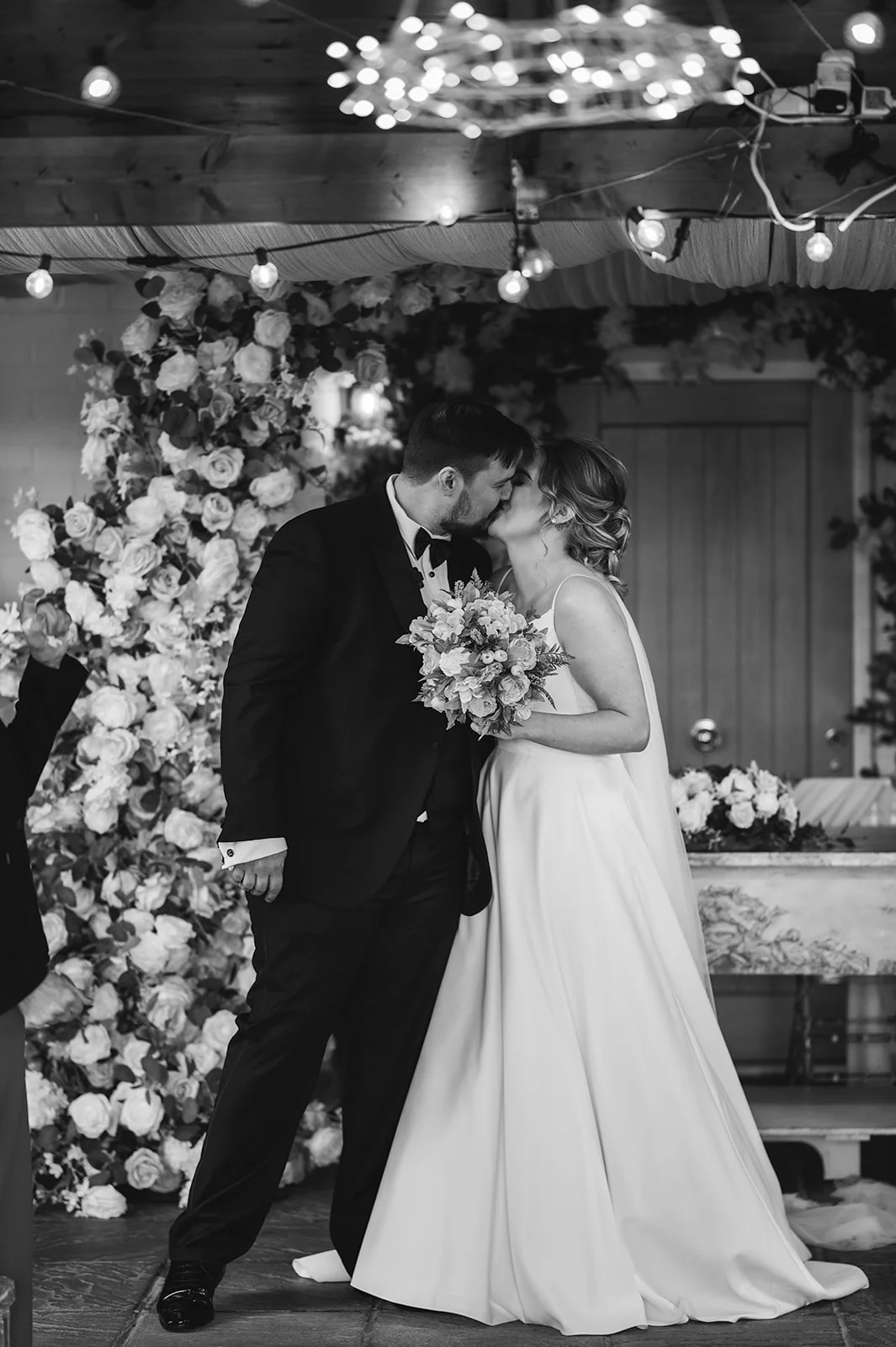 A newlywed couple shares their first kiss beneath a magnificent floral arch adorned with roses and romantic overhead lighting.