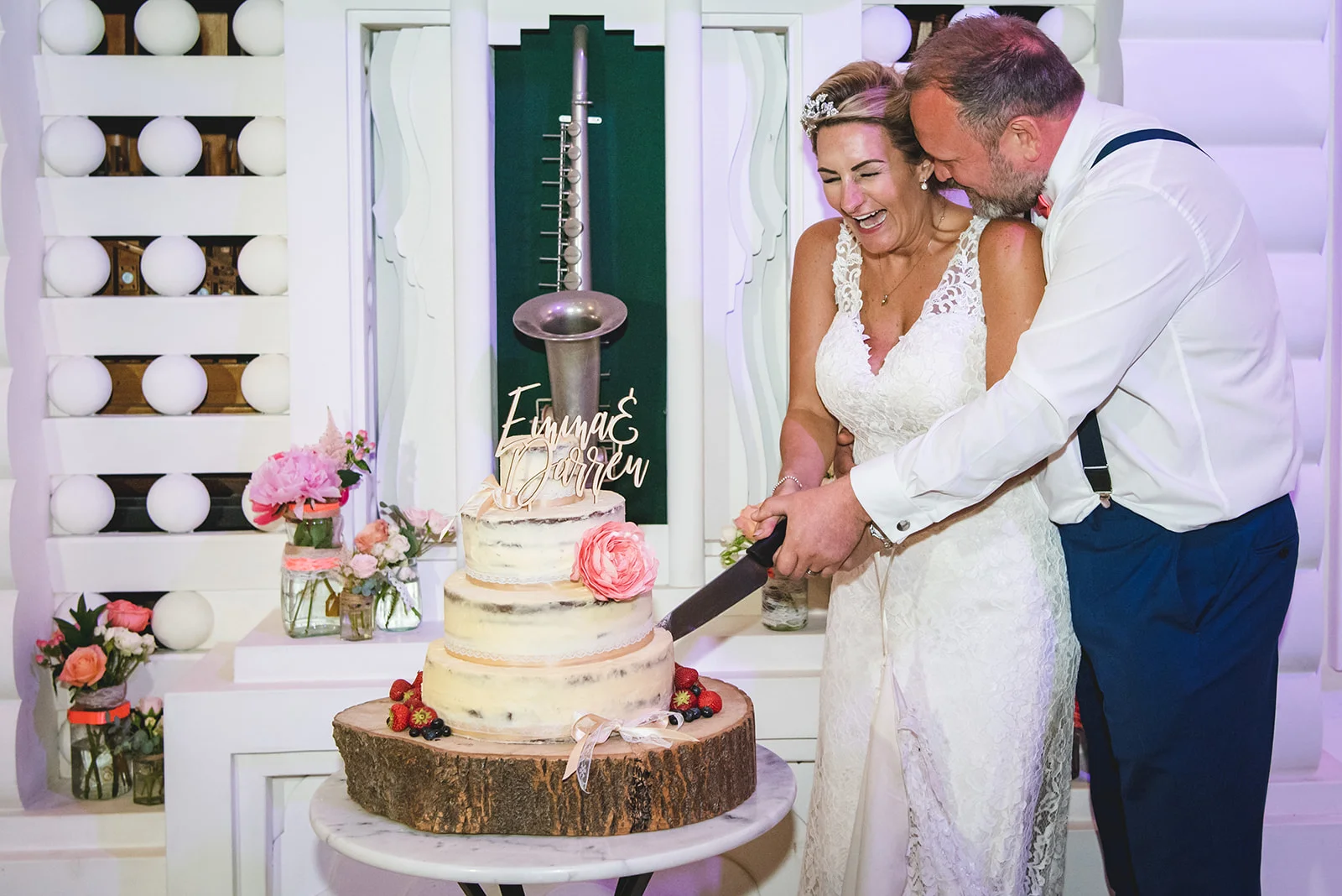 A joyful bride in a lace wedding dress and a groom in shirt sleeves laugh together as they cut their semi naked wedding cake.