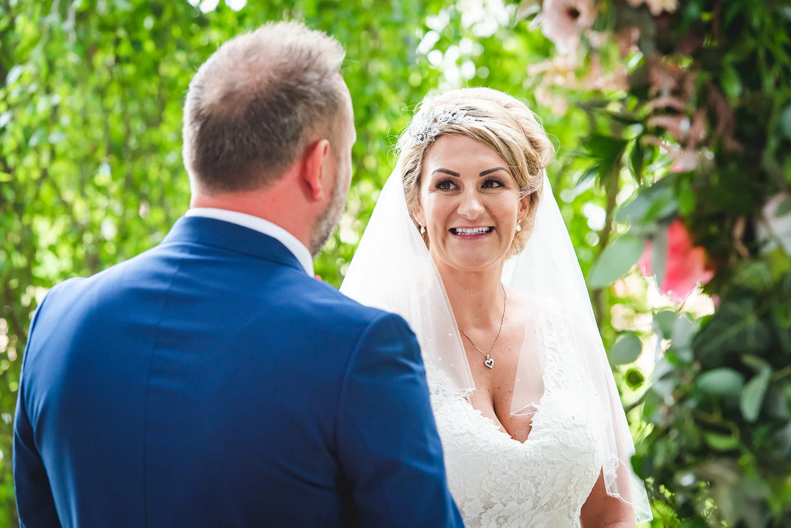 A groom in a blue suit looks lovingly at his smiling bride wearing a lace gown, veil, and tiara during an outdoor ceremony framed by green foliage.
