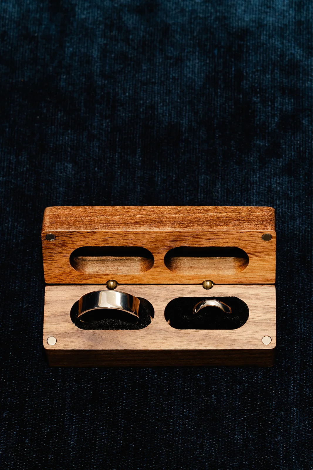 A close-up detail shot of wedding bands resting neatly inside a custom wooden box set against a dark textured surface.