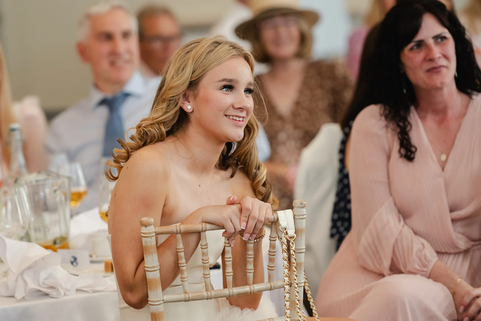 A young blonde woman smiles joyfully while seated among guests during the wedding speeches, leaning forward with her hands resting on the back of a wooden chair.