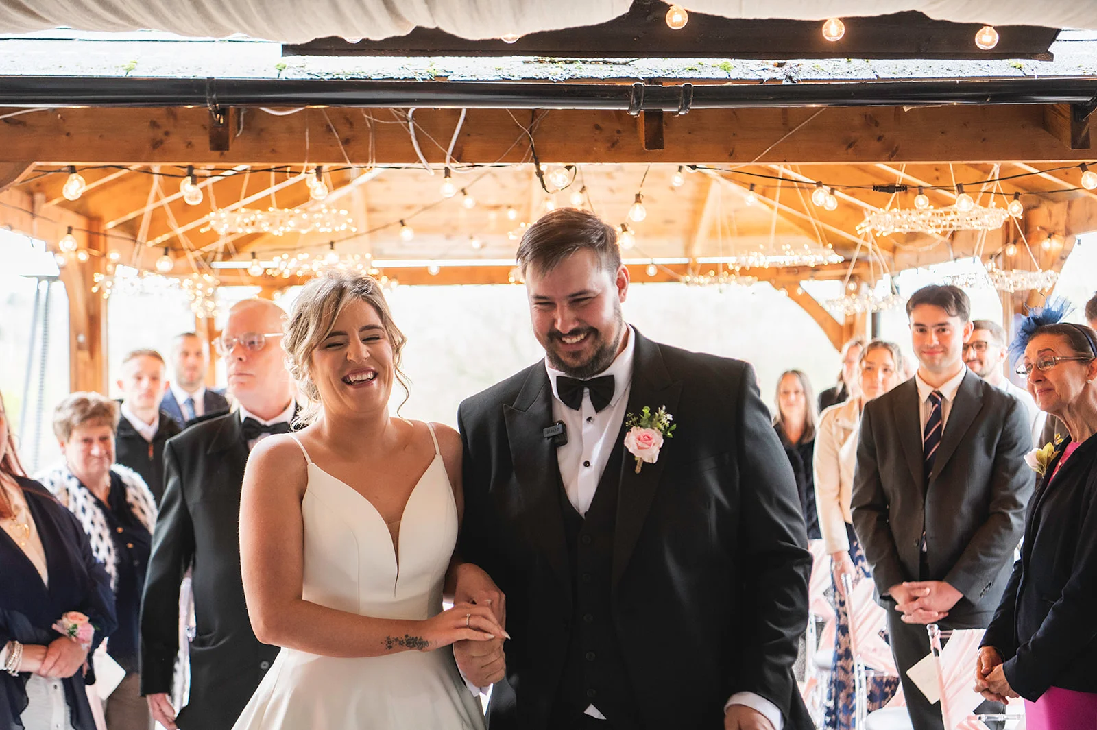 A joyful newlywed couple walks up the aisle laughing together under an oak-framed pavilion adorned with glowing fairy lights, surrounded by smiling guests.