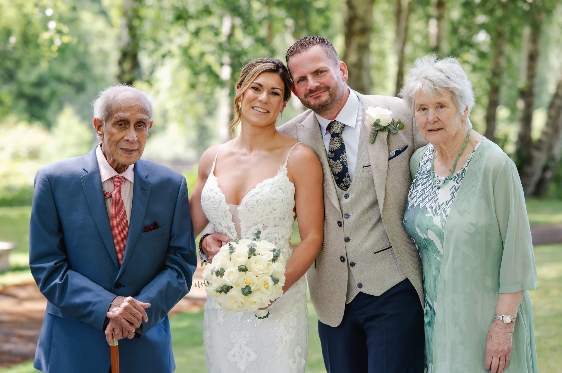 A newlywed couple poses proudly outdoors with their elderly grandparents, surrounded by leafy trees on a bright sunny day.