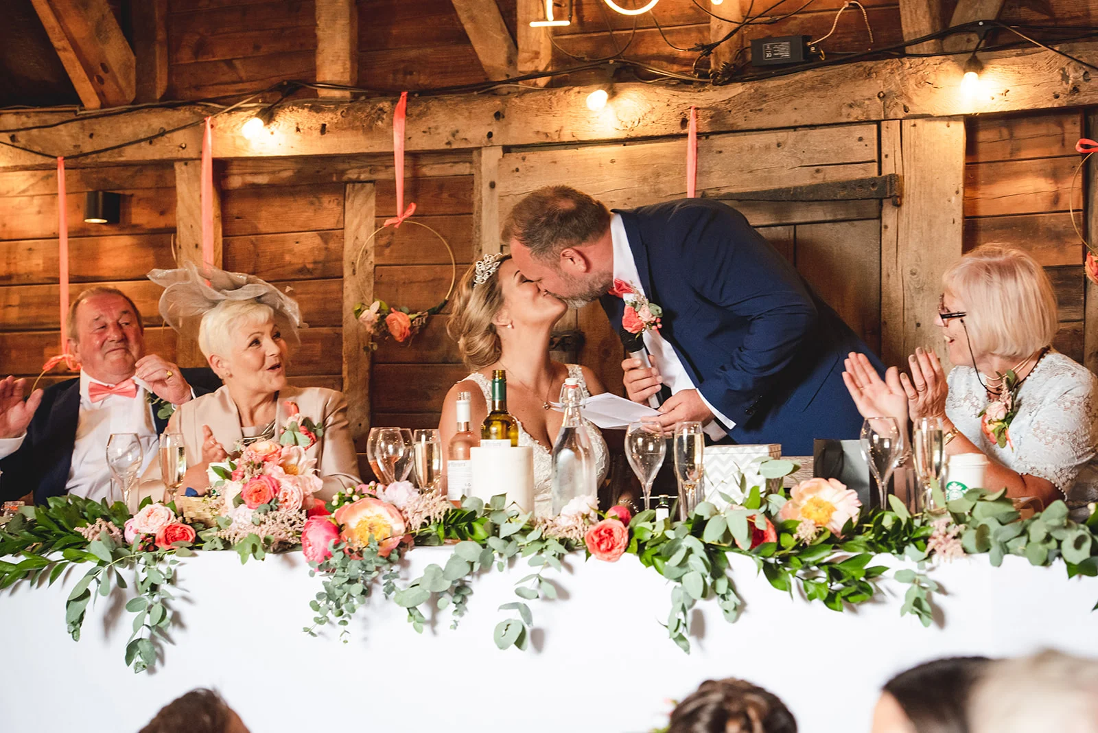 The groom leans over to kiss the bride during speeches at the top table inside a rustic wood-panelled barn decorated with floral arrangements.