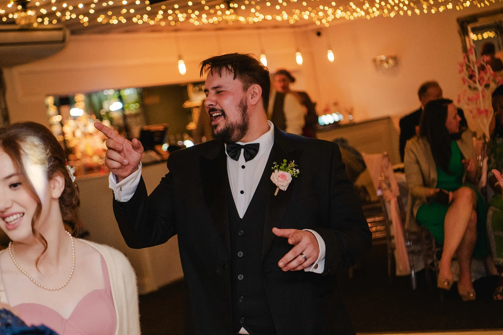 A groom in a black tuxedo and bow tie joyfully points and dances on the reception dance floor beneath a canopy of fairy lights.