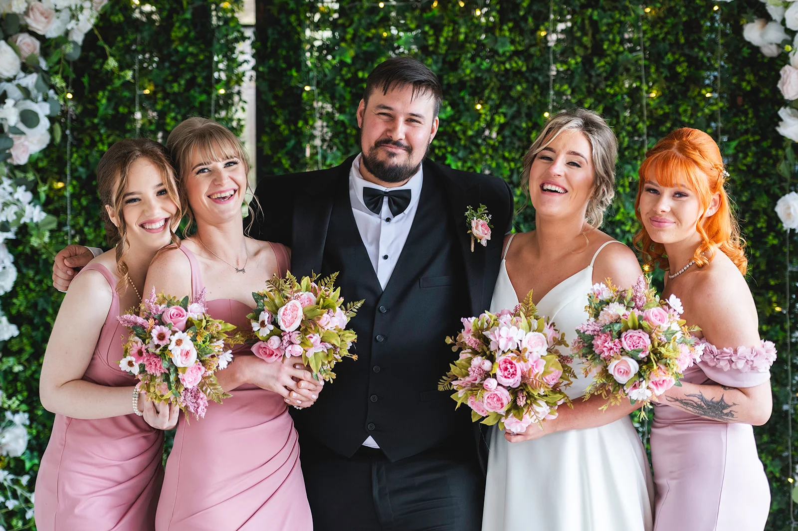 A groom in a black tuxedo stands smiling surrounded by four bridesmaids and a bride holding bouquets in front of a lush greenery wall with fairy lights.