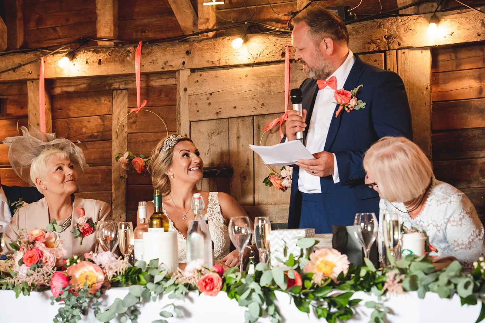The groom delivers a speech at the top table inside a rustic wood-panelled barn, while the smiling bride looks up at him beside floral arrangements.
