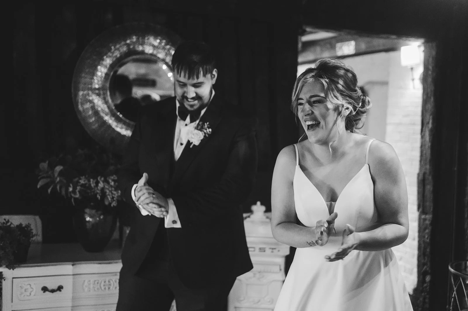 A joyful newlywed couple in black and white laughing and clapping together indoors near a decorative mirror and console table.