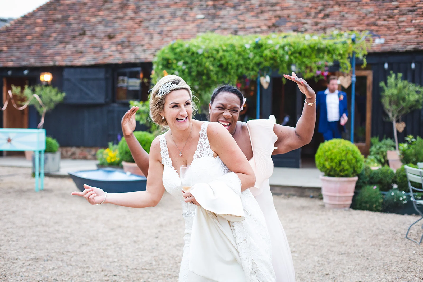 Two smiling brides share a joyful and playful moment laughing together in the gravel courtyard outside a rustic black timber wedding barn.