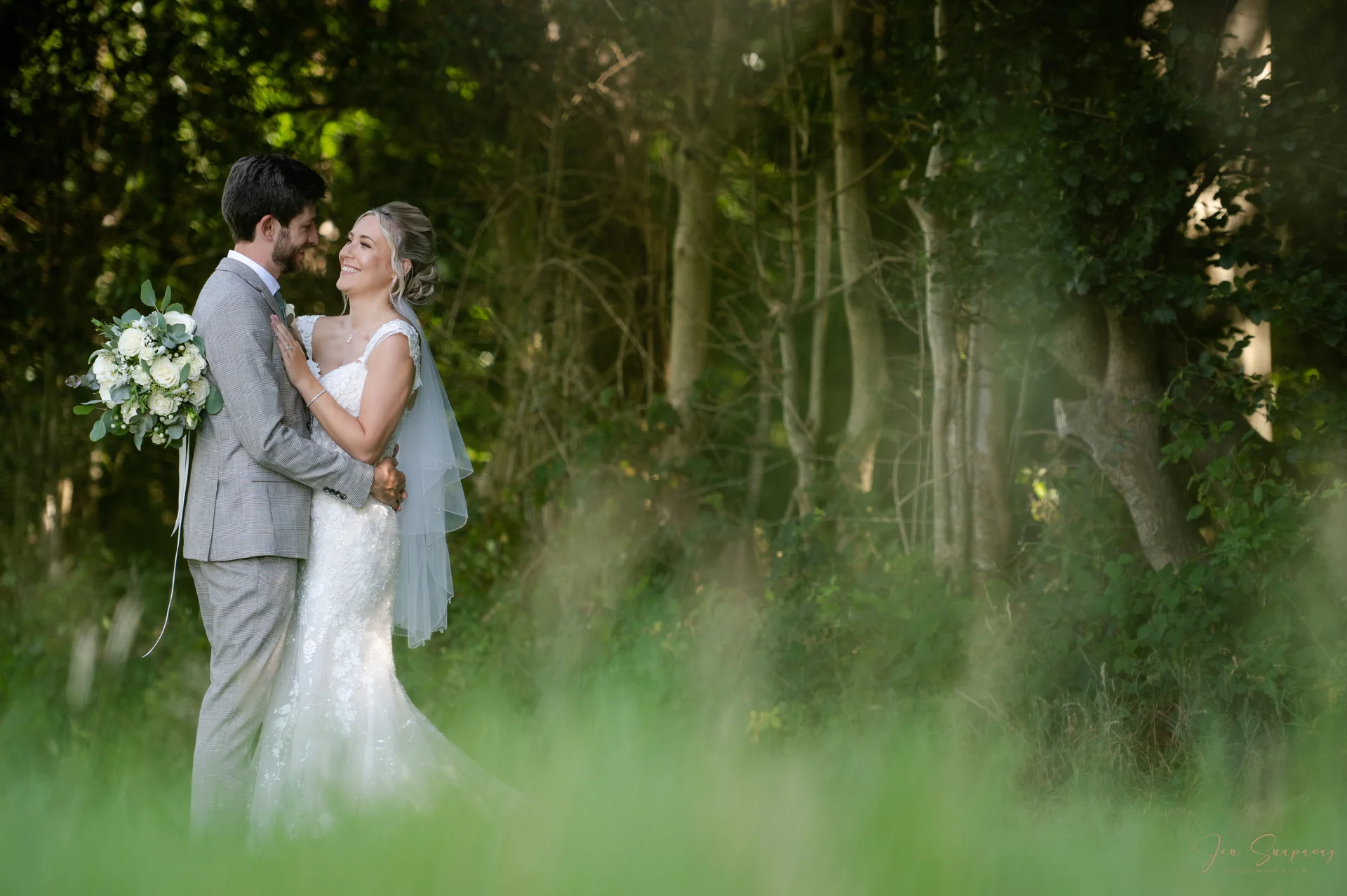 A newlywed couple embraces and smiles happily in a lush woodland setting, framed by out-of-focus foliage in the foreground.