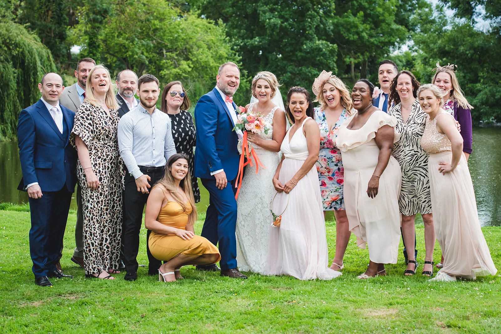 A joyful wedding group poses and laughs together outdoors on a lush green lawn beside a calm lake during a summer celebration.