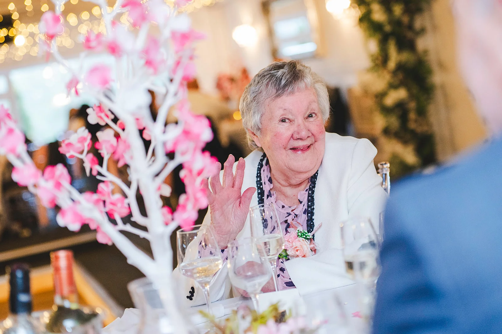 A joyful elderly guest waves cheerfully from her seat at a wedding reception table adorned with delicate pink blossom centerpieces and wine glasses.