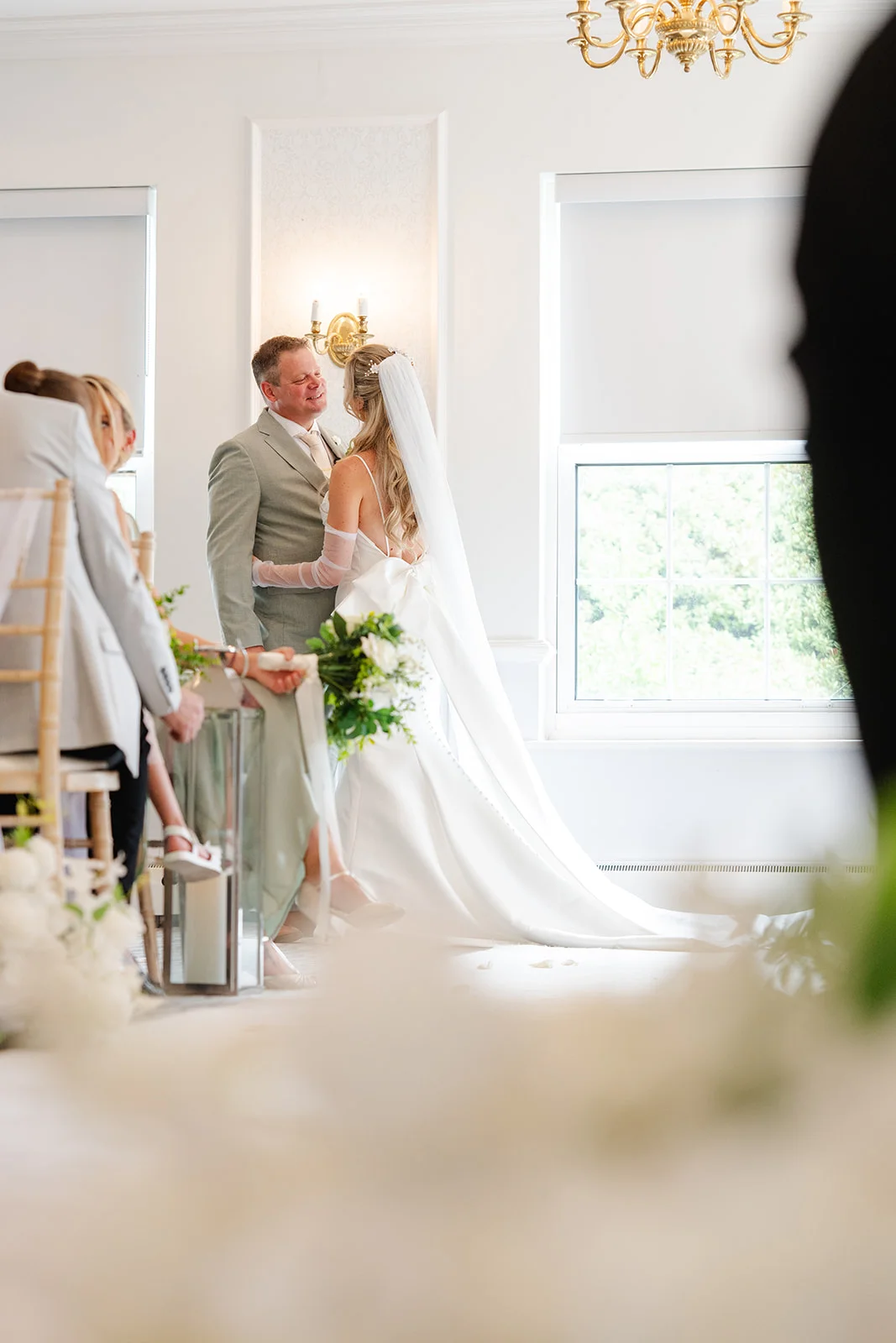 A bride and groom exchange a tender look during their indoor civil ceremony in a bright, elegant room with white walls and natural window light.
