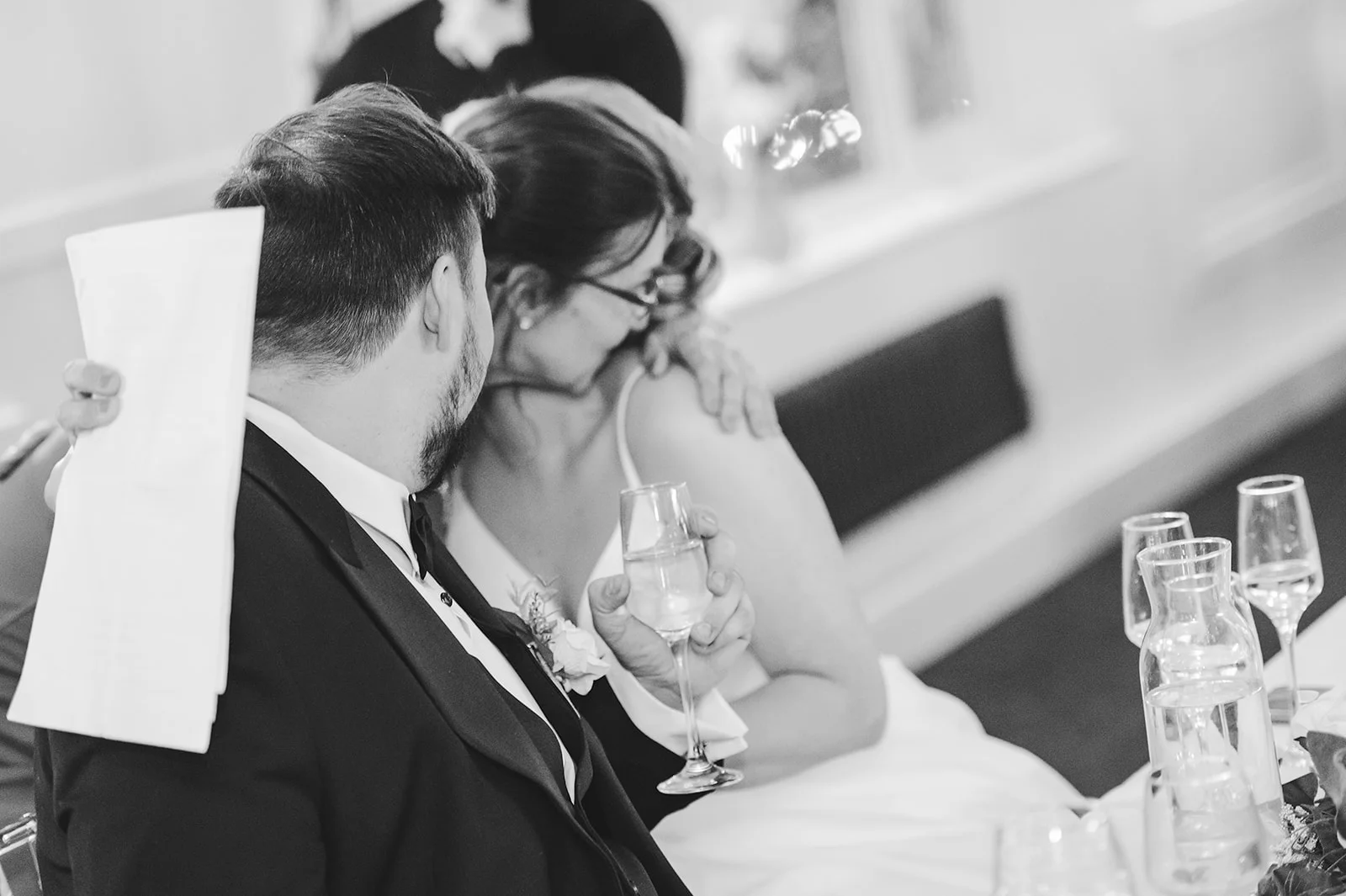 A black-and-white, over-the-shoulder shot captures a newlywed couple sharing an emotional embrace at the top table during the wedding reception.