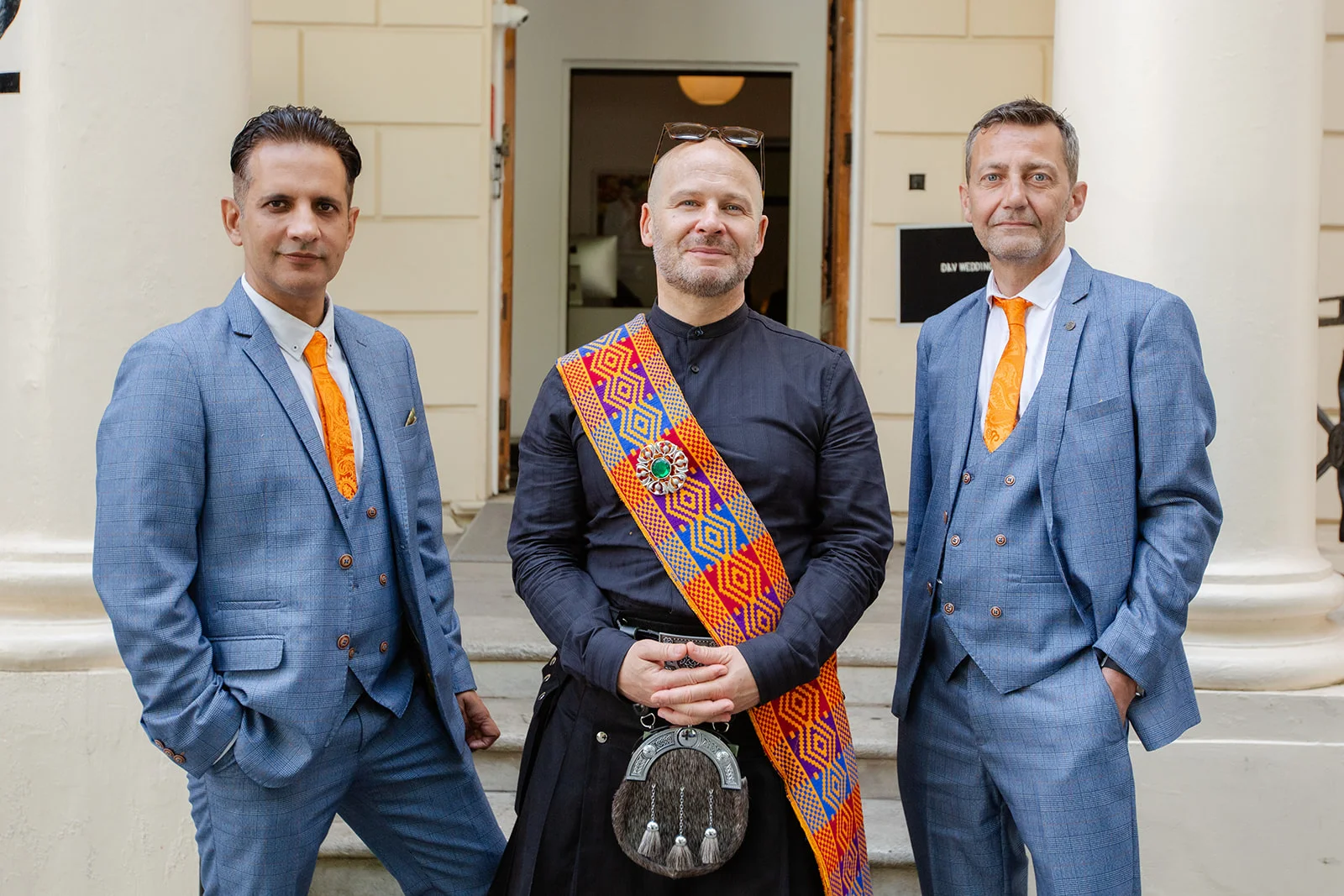 Three men pose outside a venue wearing blue checked suits with orange ties flanking a central figure in a traditional kilt and ceremonial sash.