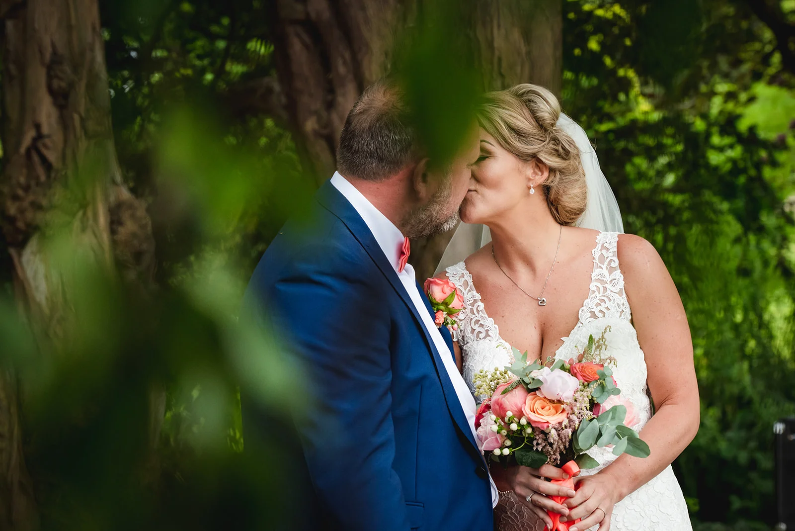A newlywed couple shares a tender kiss in a lush woodland setting, framed by green foliage with the bride holding a colourful pastel bouquet.