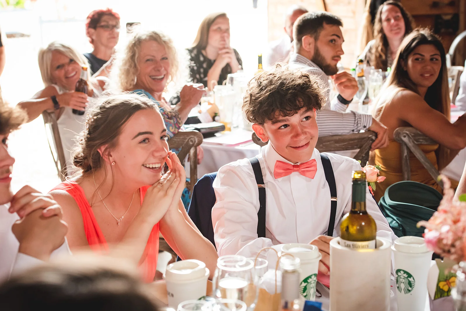 A young page boy in a white shirt, braces and a coral bow tie sits alongside smiling guests during wedding speeches, laughing joyfully.