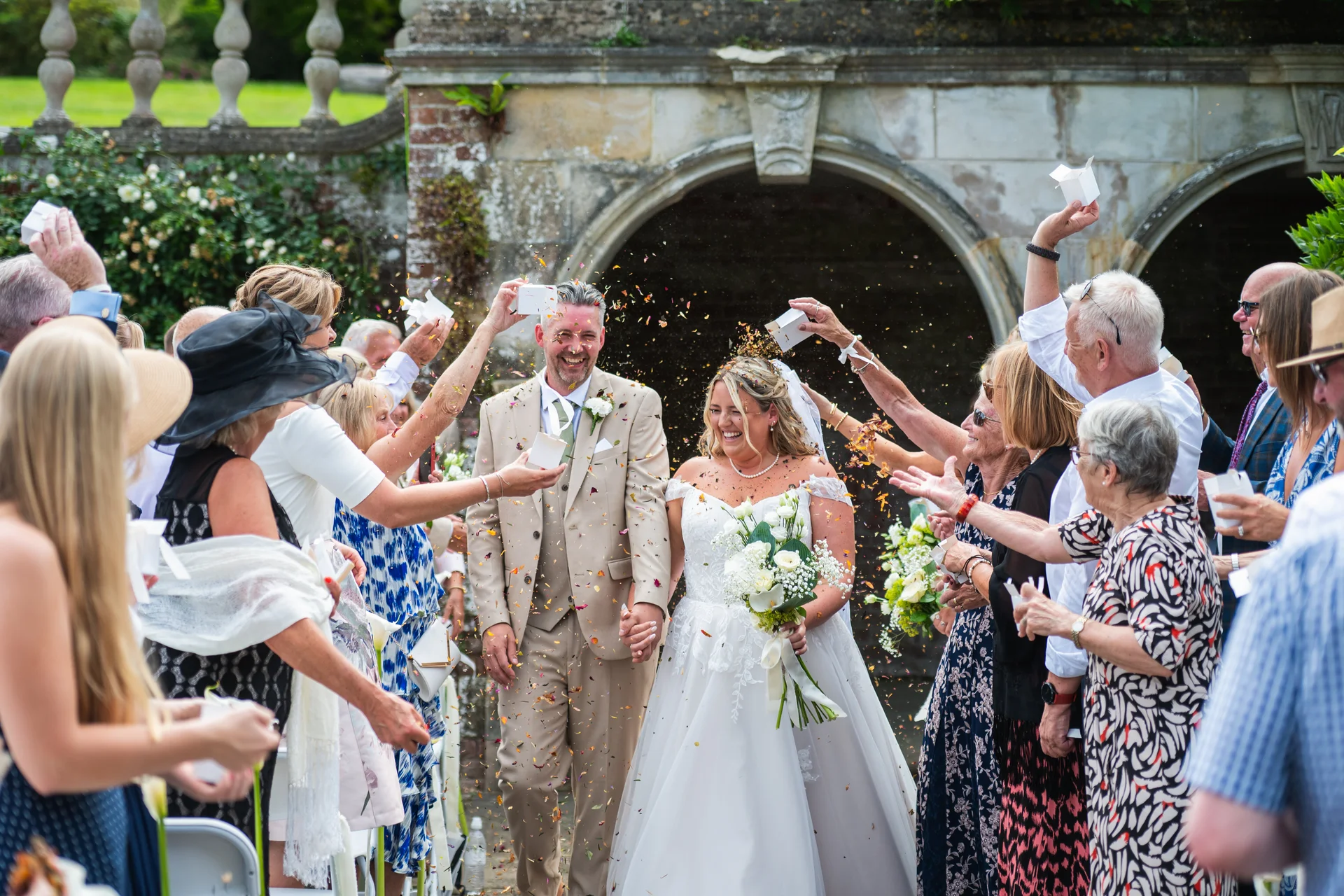 A joyful newlywed couple walks hand in hand through an enthusiastic crowd of guests throwing confetti outside a historic stone archway.
