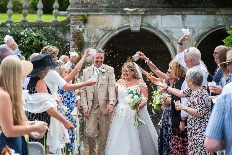 A joyful newlywed couple walks hand in hand through an enthusiastic crowd of guests throwing confetti outside a historic stone archway.