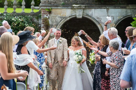 A joyful newlywed couple walks hand in hand through an enthusiastic crowd of guests throwing confetti outside a historic stone archway.