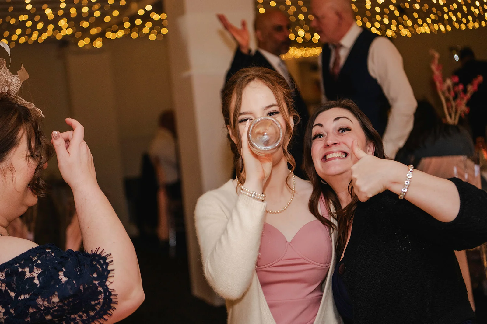 Wedding guests enjoy the evening reception under a canopy of fairy lights, with one woman drinking from a glass and another giving a thumbs up.
