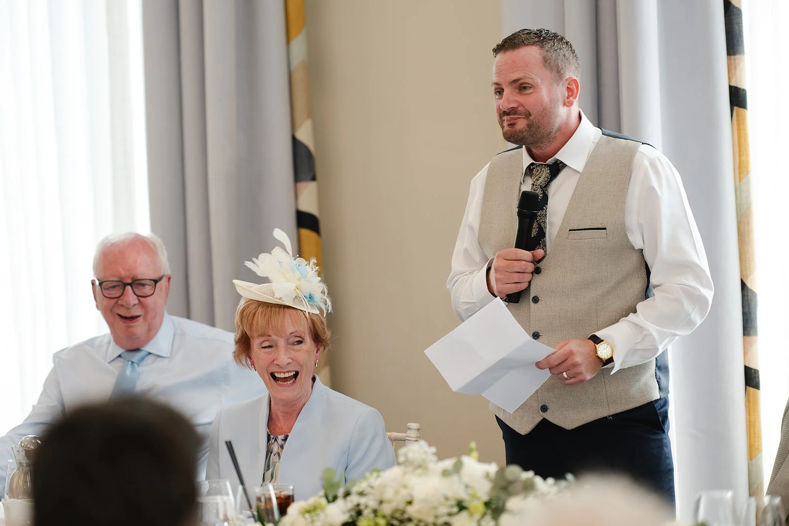 A groom delivers a speech holding a microphone and notes, while seated wedding guests including a woman wearing a fascinator laugh joyfully during the wedding breakfast.