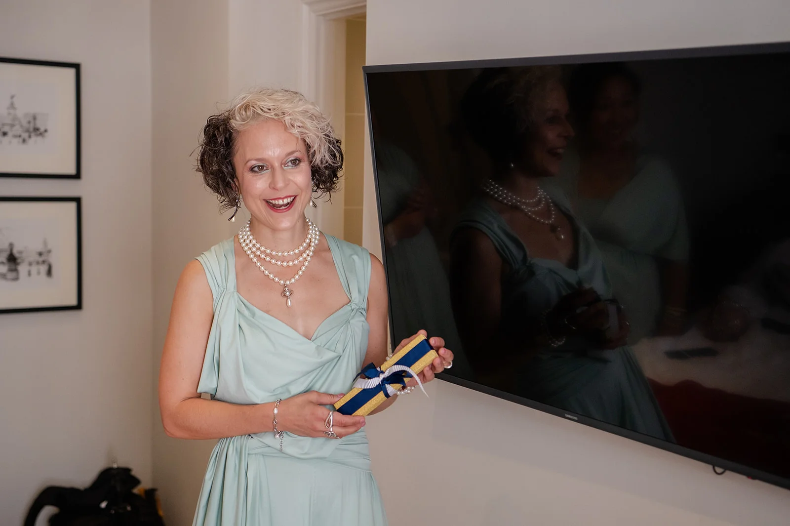 A smiling woman wearing a sage green draped gown and layered pearl necklaces holds a gift box during wedding preparations, reflected in a large screen behind her.