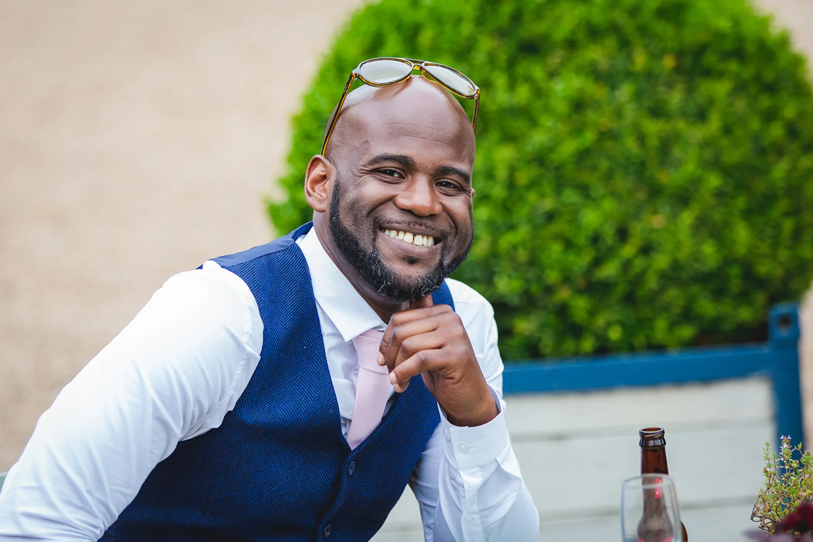 A smiling groom wearing a blue waistcoat and pink tie relaxes outdoors with sunglasses resting on his head.